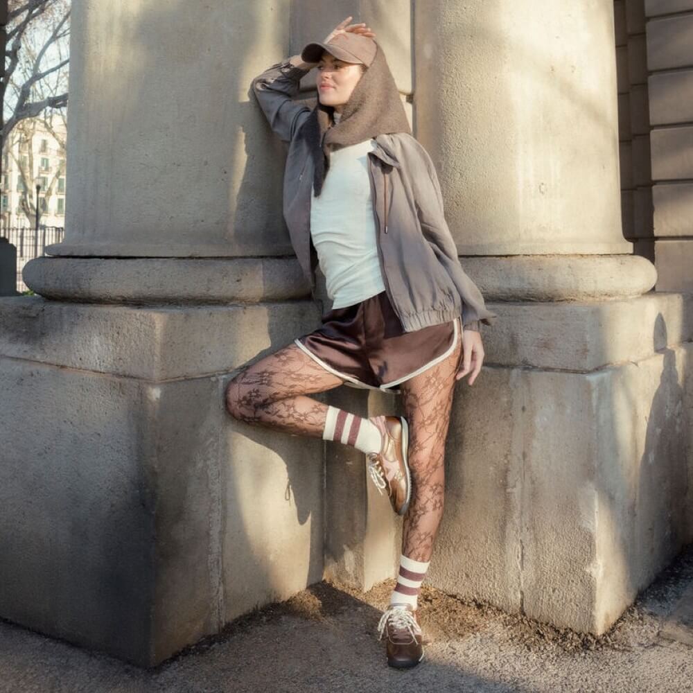 A model wearing a brown cap, brown hooded jacket, white tank top, brown satin skirt, patterned tights, and Victoria brown sneakers leans against a large stone column outdoors.