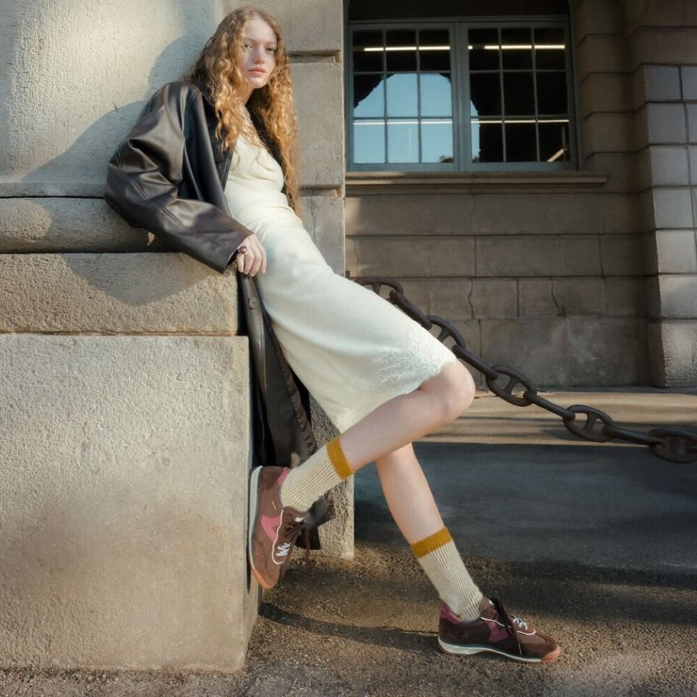 A model leans against a stone wall wearing a cream-colored textured dress, a black leather jacket, cream socks with yellow stripes, and brown Victoria lace-up shoes, with a large black chain running behind her.