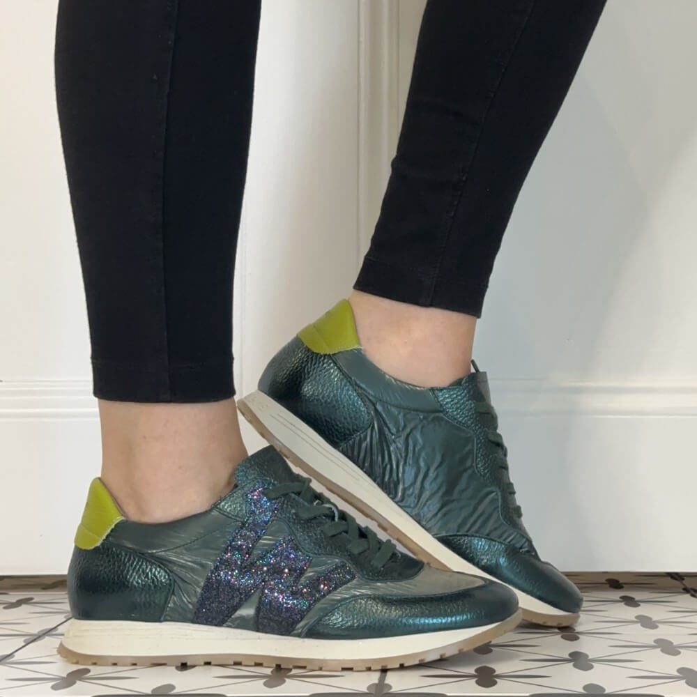 A person wearing Wonders dark green textured leather sneakers with a glittery purple side stripe, paired with black leggings, standing on a patterned tile floor against a white wall.