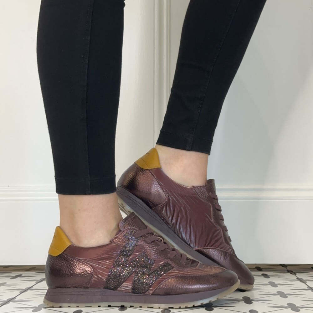 A person wearing black leggings and Wonders wine textured sneakers with yellow heel accents, standing on a patterned tile floor against a white wall.