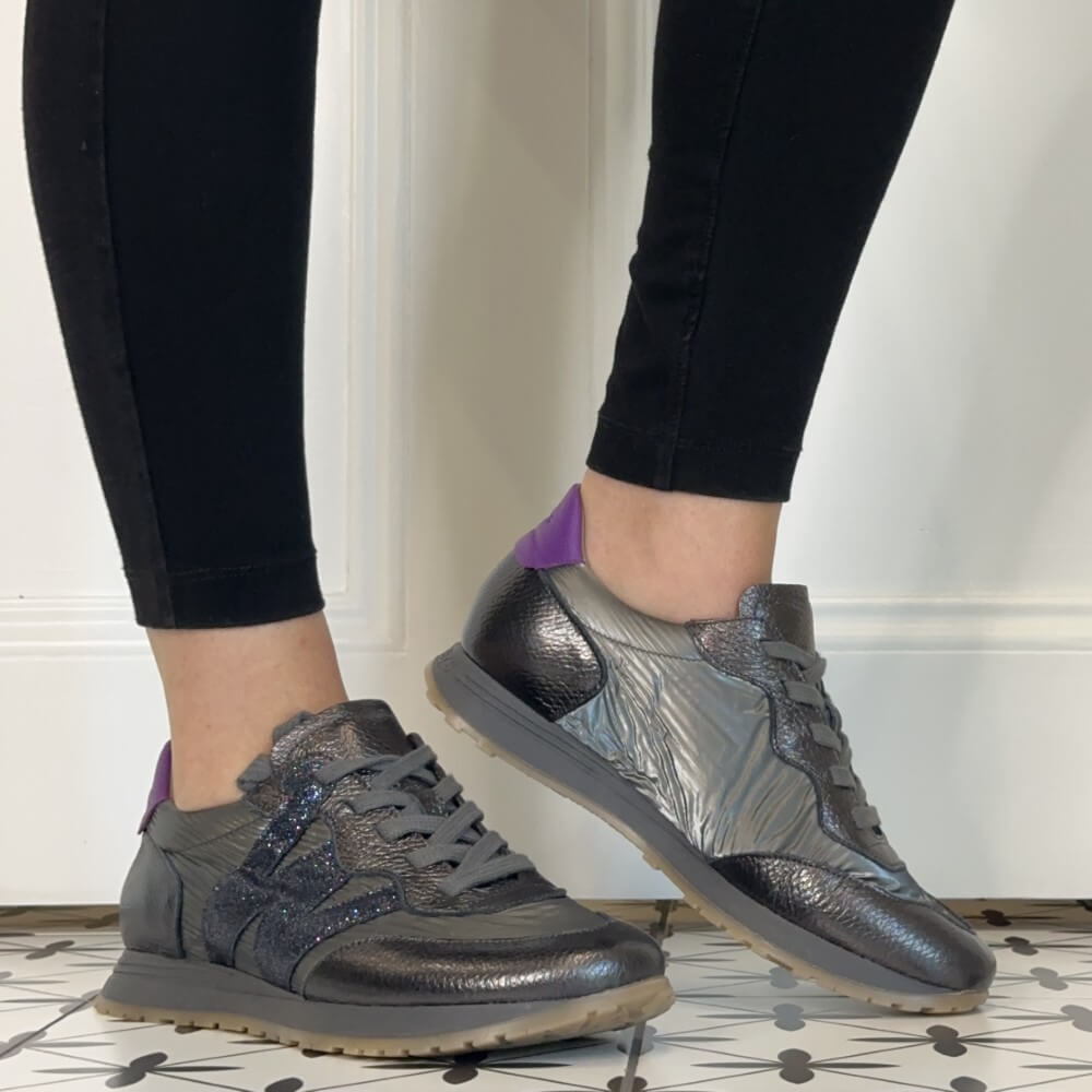 A person wearing Wonders metallic grey and silver sneakers with purple heel tabs, shown from the calves down against a white panelled wall and patterned tile floor.