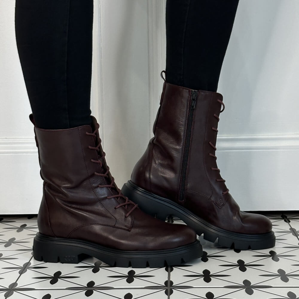 A pair of Wonders Wine leather lace-up combat boots with side zippers and chunky black soles, shown on a person wearing black leggings standing on a black-and-white patterned tile floor.