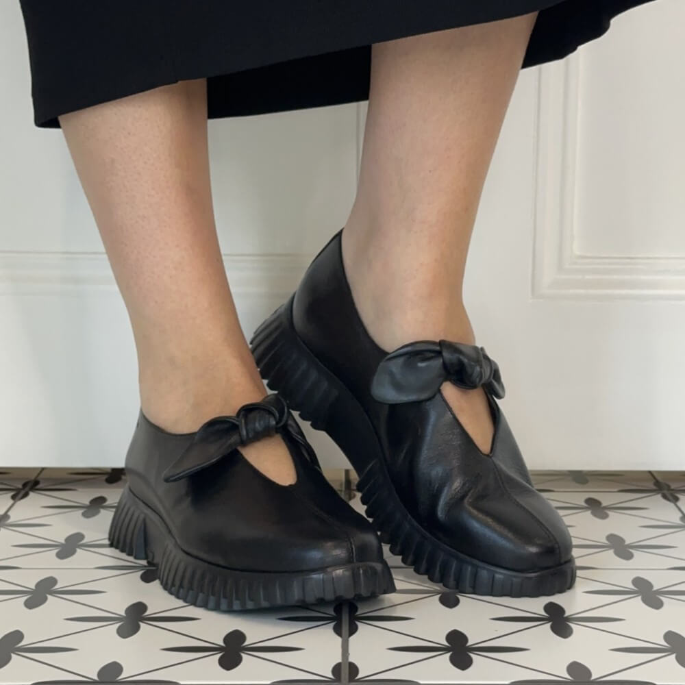 Wonders Black leather Mary Jane loafers with chunky lug soles and bow details, shown on a person's feet standing on a patterned tile floor.