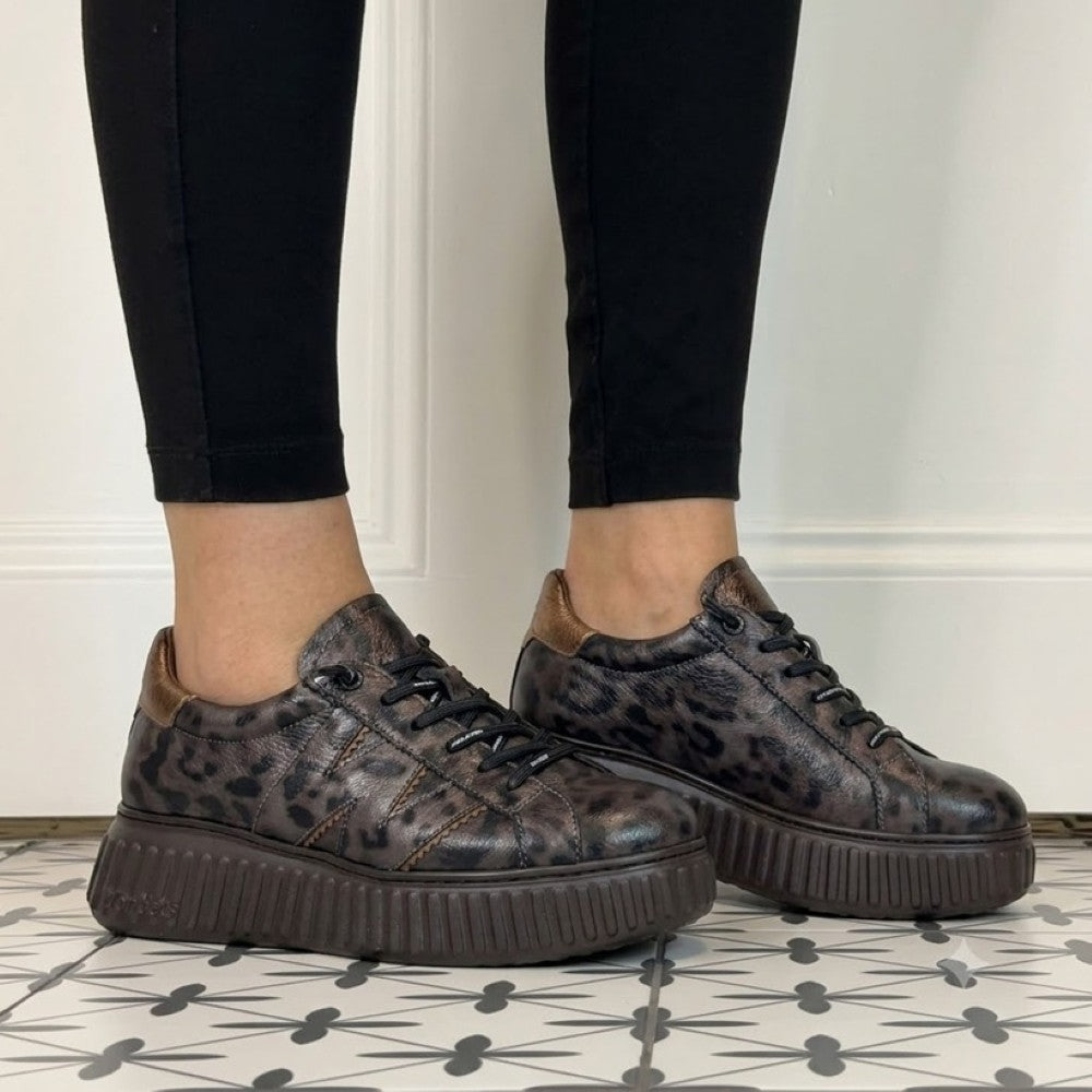 A person wearing black leggings and Wonders dark brown platform sneakers with a distressed leopard print pattern, standing on a black and white patterned tile floor.