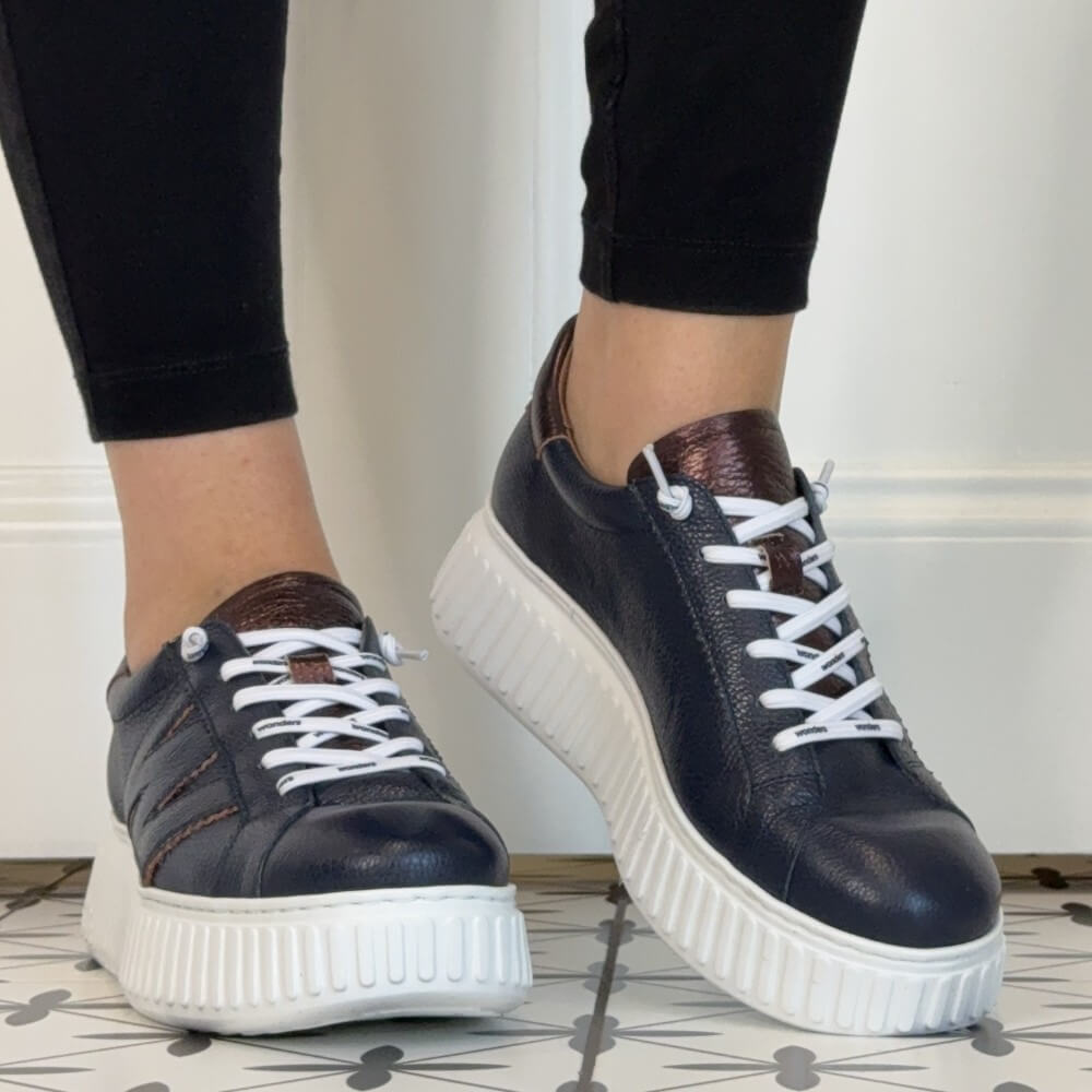 A pair of Wonders low-top platform sneakers in dark navy blue leather with white laces and thick white platform soles, shown being worn with black leggings.