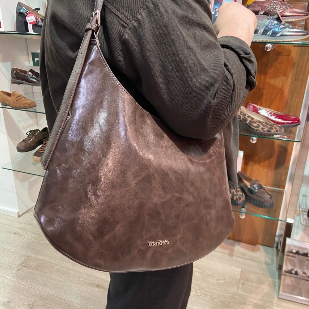 A person wearing a dark brown jacket carries a Wonders large, slouchy brown leather shoulder bag with a distressed finish. The brand name 'Wonders' is printed in small gold letters near the bottom of the bag.