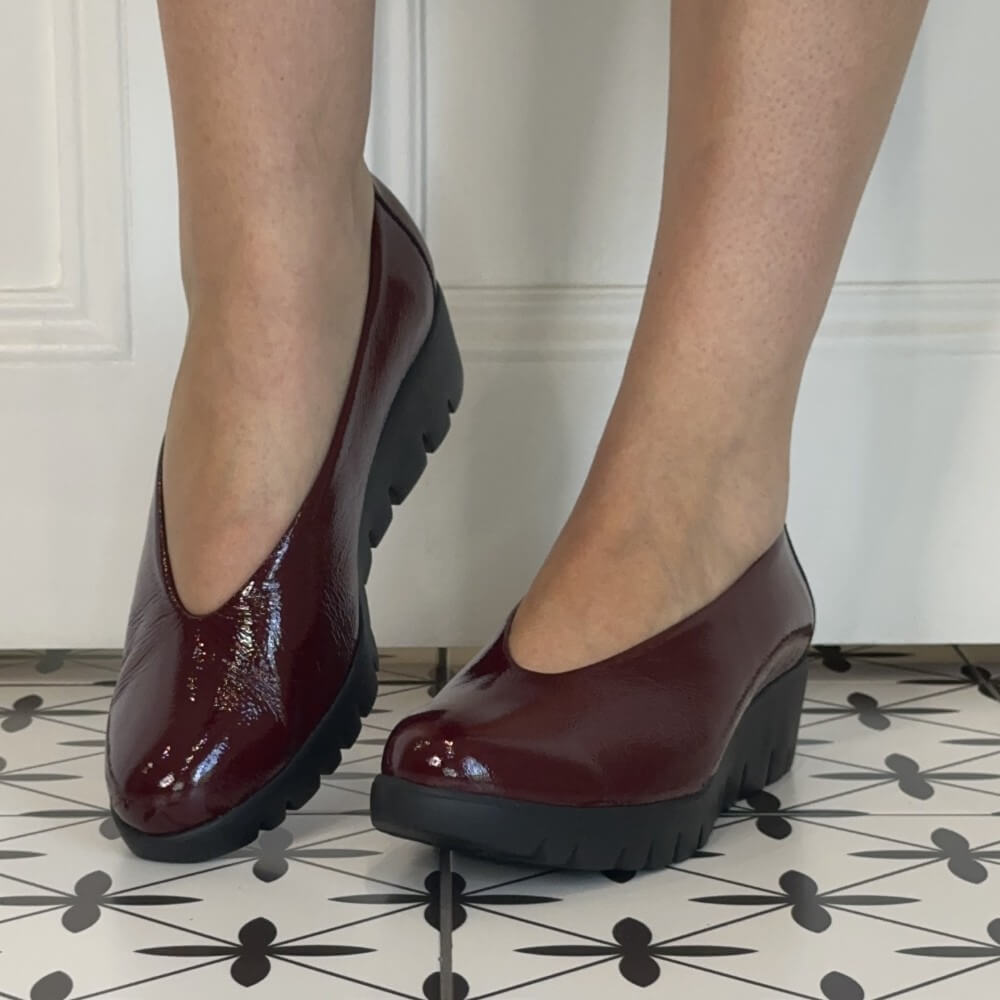 A pair of Wonders glossy burgundy platform wedge pumps with black lug soles, shown on a person's feet against a white panelled wall and patterned tile floor.