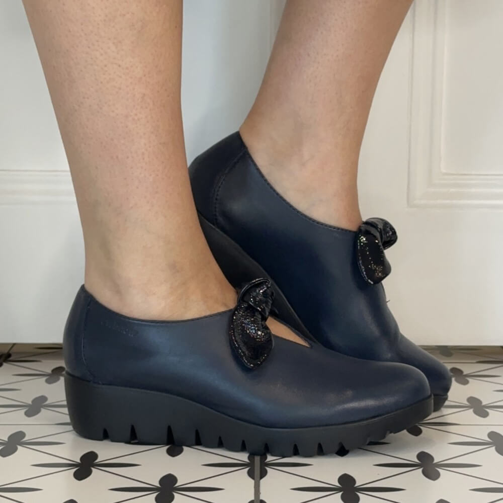 Wonders navy leather slip-on with a patent bow trim and a black wedge sole worn on a tiled floor