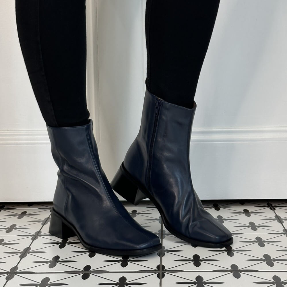 A pair of Wonders dark navy blue leather ankle boots with low block heels, worn with black leggings on a black and white patterned tile floor.