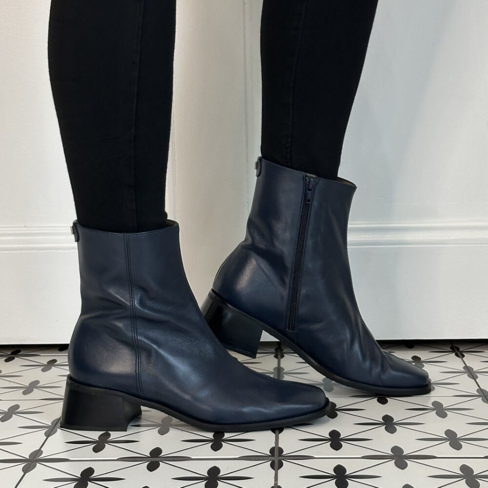 A pair of Wonders dark navy blue leather ankle boots with low block heels, worn with black leggings on a black and white patterned tile floor.