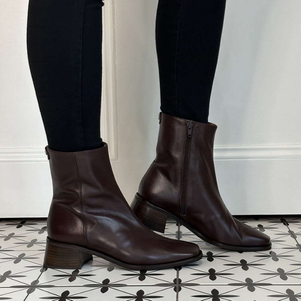 A pair of Wonders dark brown leather ankle boots with side zippers, worn with black leggings on a patterned black and white tile floor.
