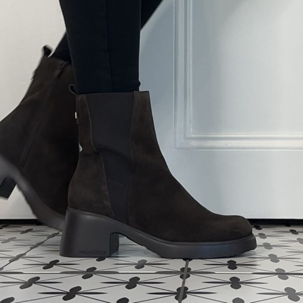 A pair of Wonders dark brown suede Chelsea-style ankle boots with elastic side panels and chunky black block heels, shown on a person wearing black leggings on a patterned floor.