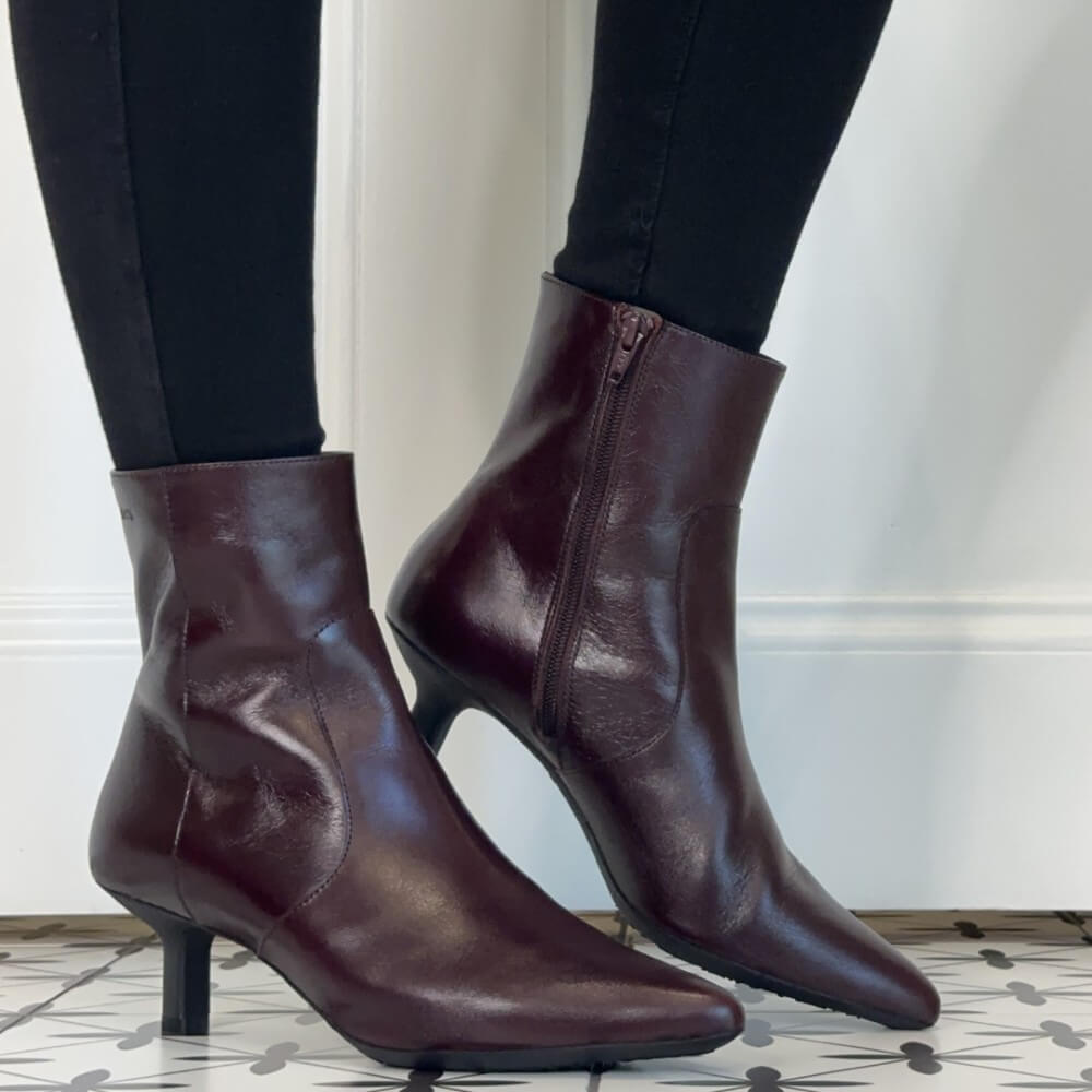 A pair of Wonders dark burgundy leather ankle boots with kitten heels, shown on a model wearing black leggings against a white wall and patterned tile floor.