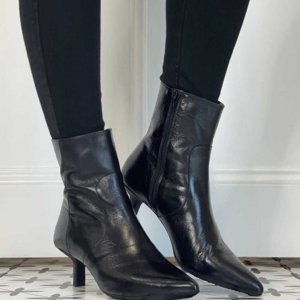A pair of Wonders black leather ankle boots with pointed toes and mid-height stiletto heels, worn with black leggings. The boots are shown from the side and back, highlighting the side zipper and leather texture, set against a white wall and patterned tile floor.