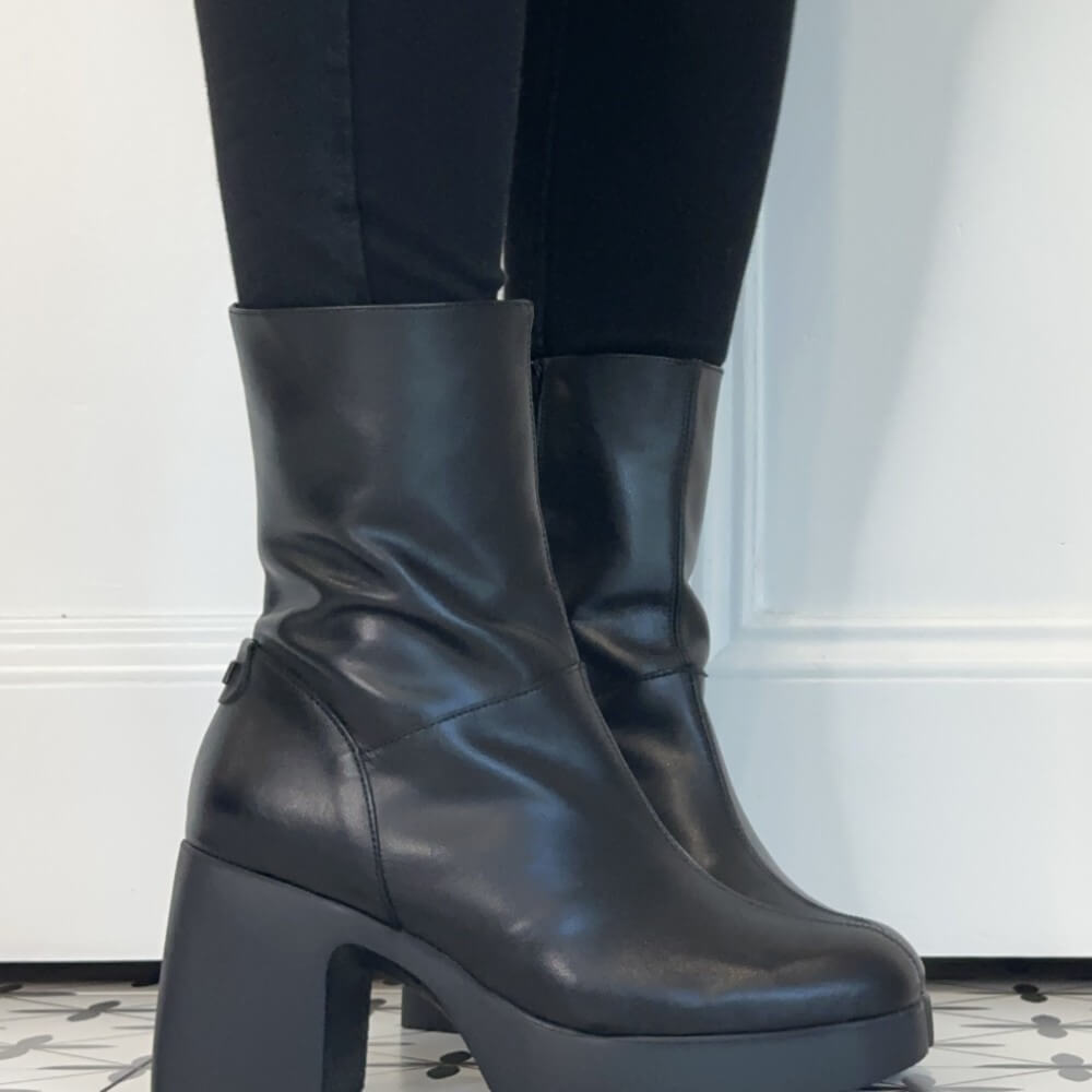 Wonders Black leather ankle boots with chunky block heels and platform soles, shown worn with black leggings against a white panelled wall.