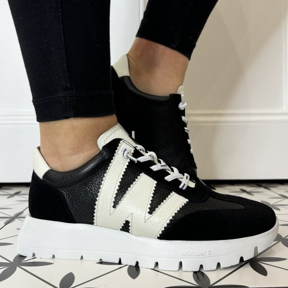 Black and white sneakers with a brand logo worn indoors on a patterned floor.