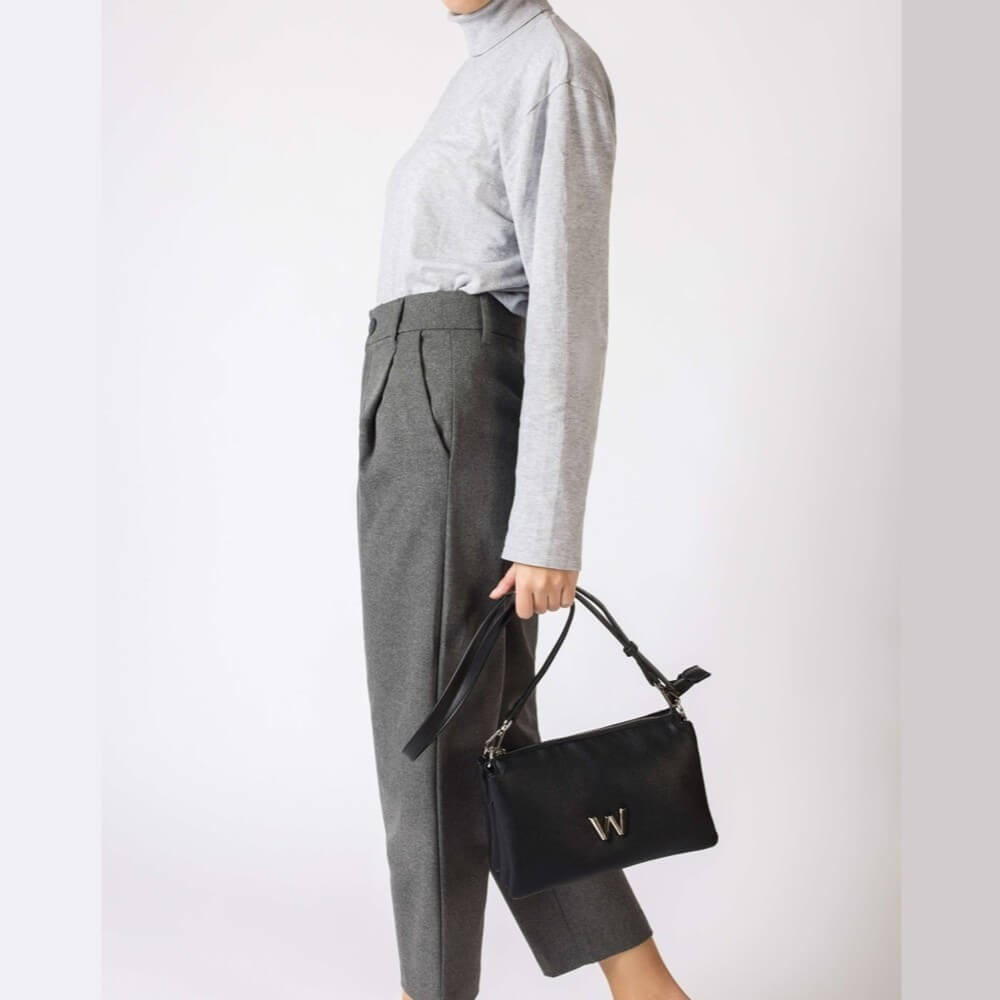 Person wearing a gray turtleneck and gray pants holding a black handbag with a visible brand logo on a white background