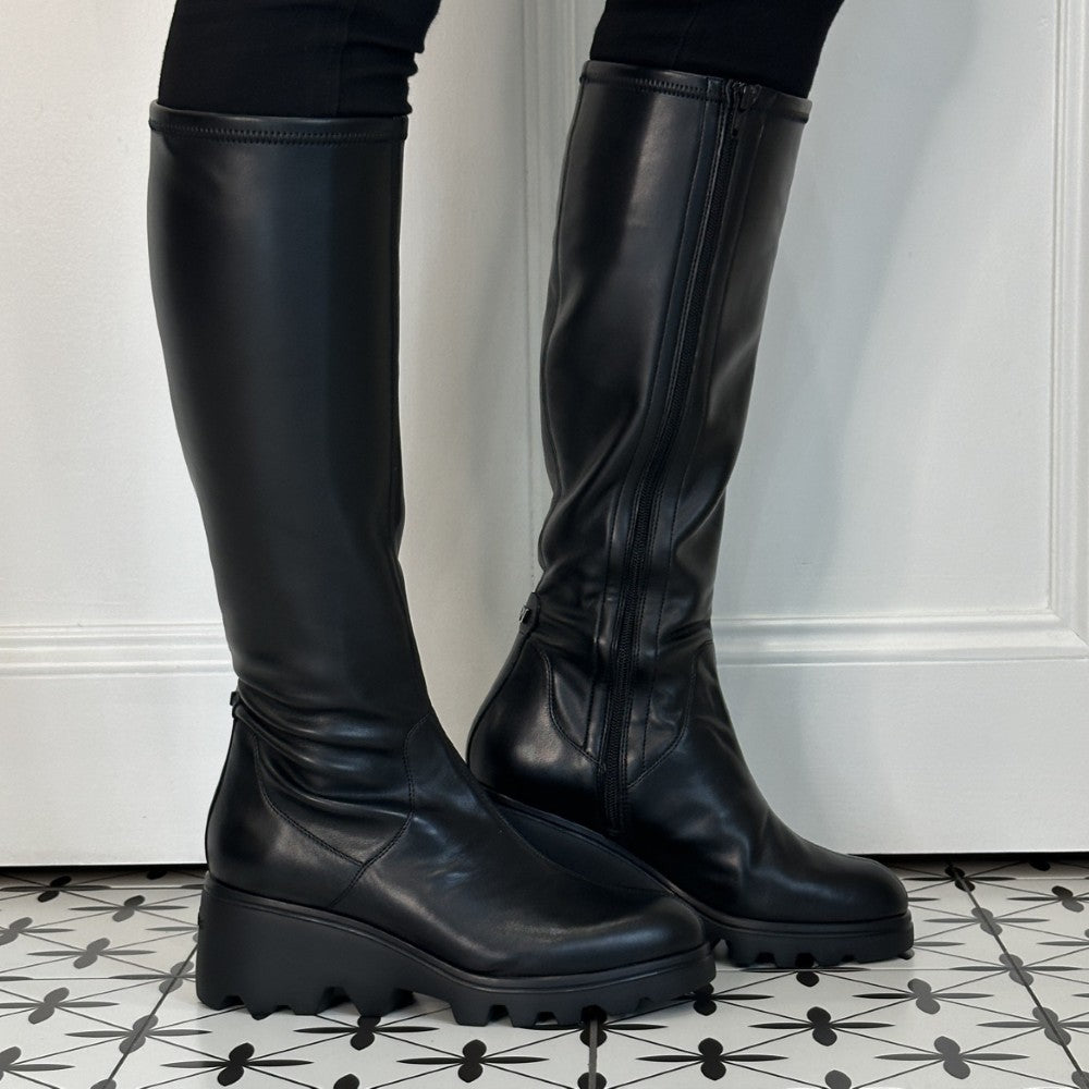 A pair of Wonders black leather knee-high boots with side zippers and chunky lug soles, shown worn with black leggings on a patterned black-and-white tile floor against a white panelled wall.