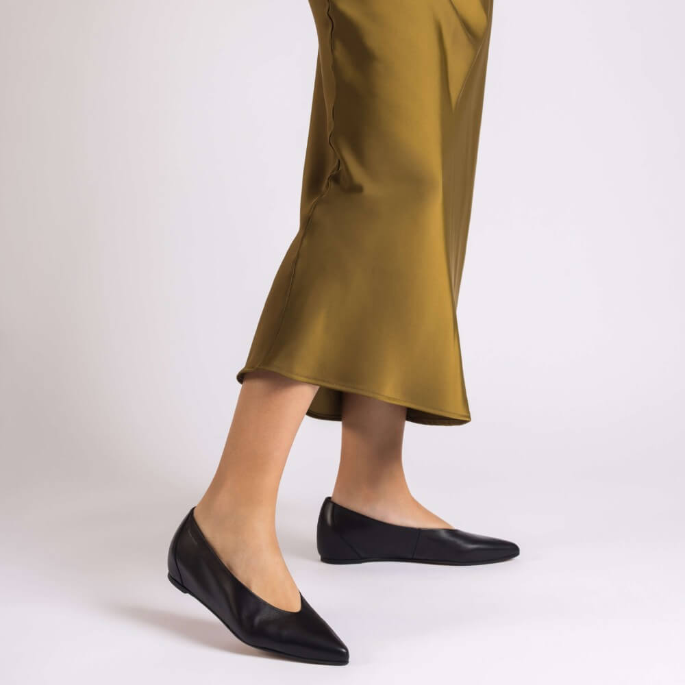 A person wearing Wonders black leather ballet flats with a pointed toe, paired with a flowing olive green satin skirt, shown from the knees down against a light grey studio background.