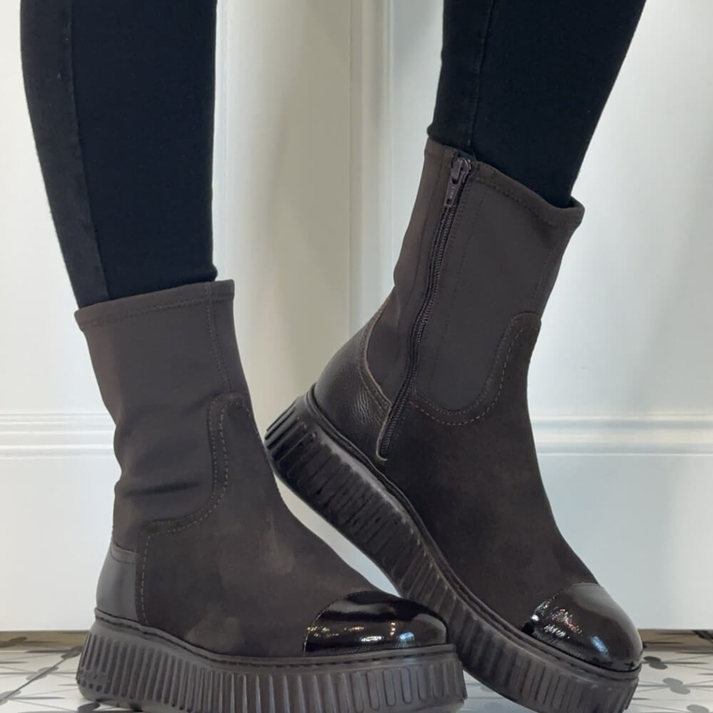A pair of Wonders dark brown ankle boots with elastic side panels, chunky lug soles, and glossy patent leather toe caps, shown on a model wearing black leggings.