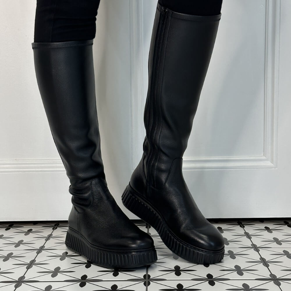 A pair of Wonders black knee-high leather boots with chunky lug soles, shown on a model's feet against a white panelled door and black-and-white patterned tile floor.