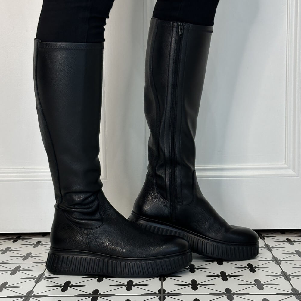 A pair of Wonders black knee-high leather boots with chunky lug soles, shown on a model's feet against a white panelled door and black-and-white patterned tile floor.