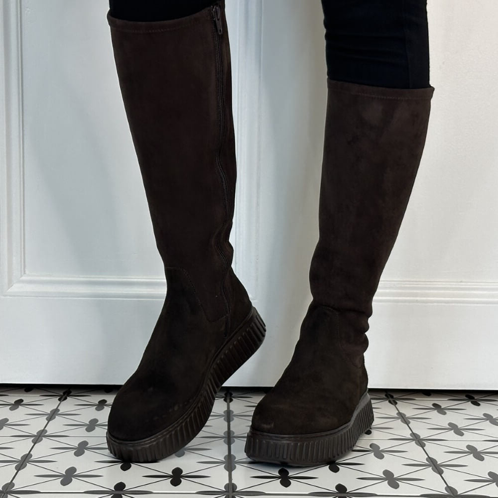 A pair of Wonders dark brown suede knee-high platform boots with side zippers, shown on a model's legs against a white panelled wall and black-and-white patterned tile floor.