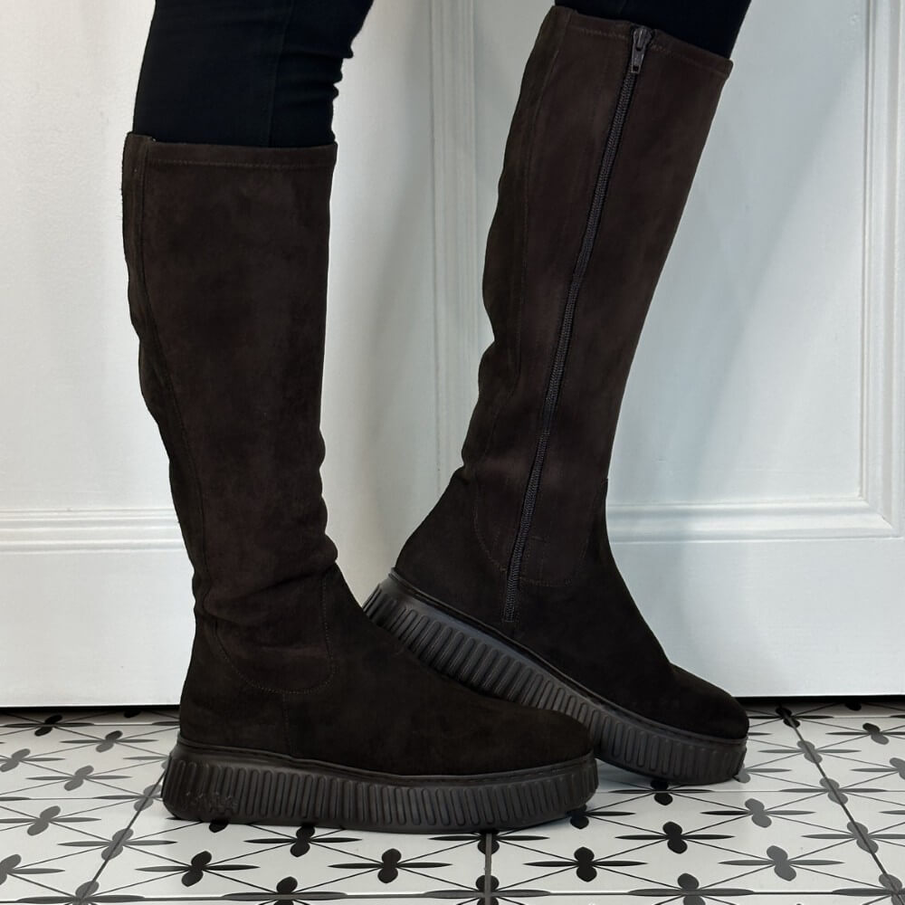 A pair of Wonders dark brown suede knee-high platform boots with side zippers, shown on a model's legs against a white panelled wall and black-and-white patterned tile floor.