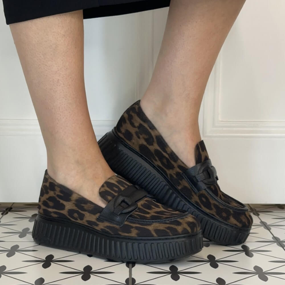 A person wearing Wonders brown and black leopard print platform loafers with black soles, standing on a black and white patterned tile floor against a white panelled wall.