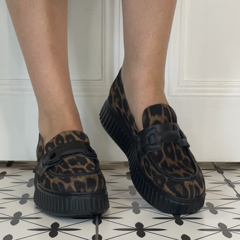 A person wearing Wonders brown and black leopard print platform loafers with black platform soles, standing on a black and white patterned tile floor in front of a white paneled wall.