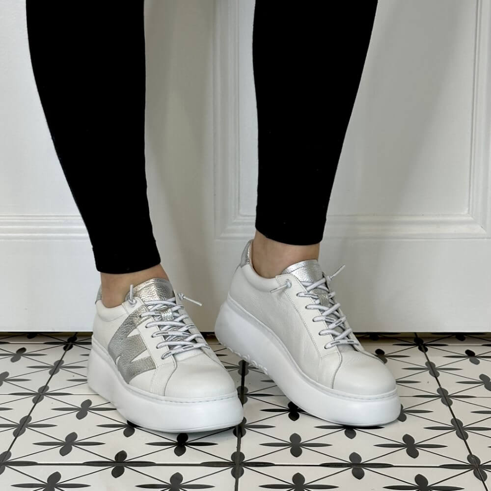 White sneakers with silver accents worn by a person on a patterned floor.