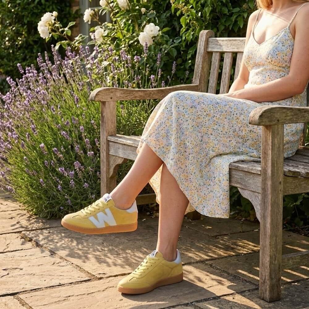 Woman sitting on a wooden bench in a garden wearing yellow sneakers with a floral dress.