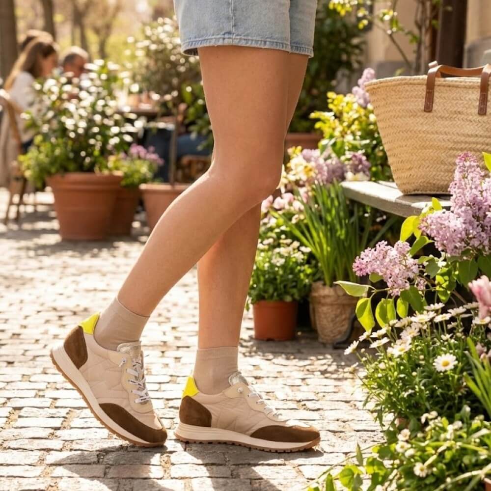 Person wearing beige sneakers with brown accents walking on a cobblestone path surrounded by flowers and plants.