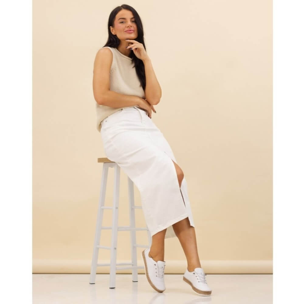 Woman sitting on a stool wearing a beige sleeveless top and white skirt against a beige background