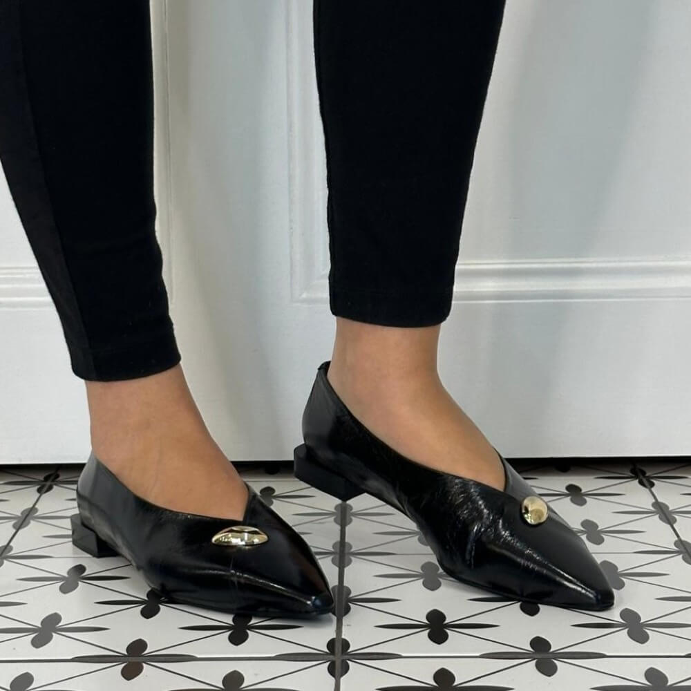 A person wearing Angel Alarcon black pointed-toe flats with small gold-tone decorative accents, shown from the calves down against a white wall and black-and-white patterned tile floor.