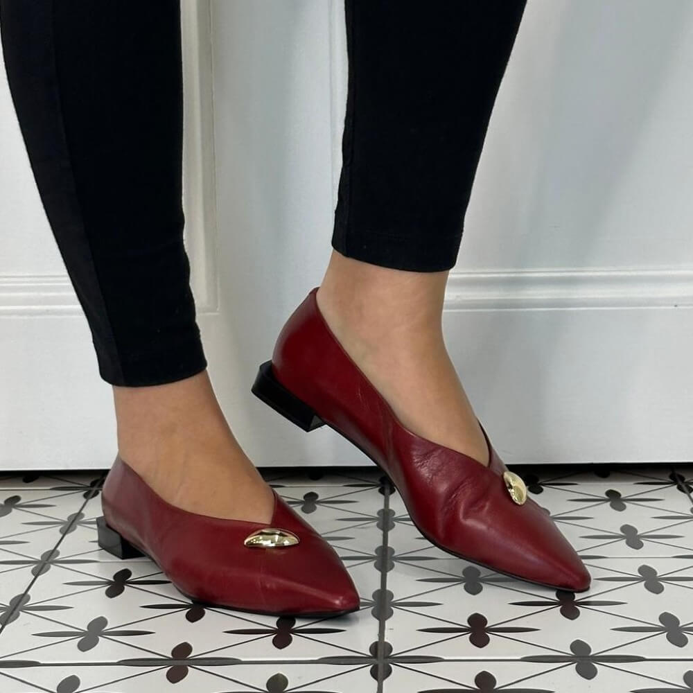 A person wearing black leggings and Angel Alarcon red leather pointed-toe flats with small gold-tone decorative accents, standing on a black-and-white patterned tile floor against a white wall.