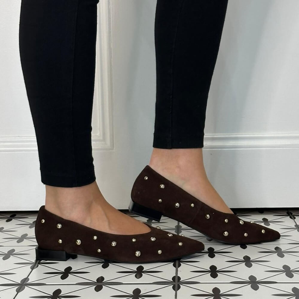 A person wearing Angel Alarcon dark brown suede pointed-toe flats with silver stud embellishments, paired with black leggings, standing on a black and white geometric patterned tile floor.