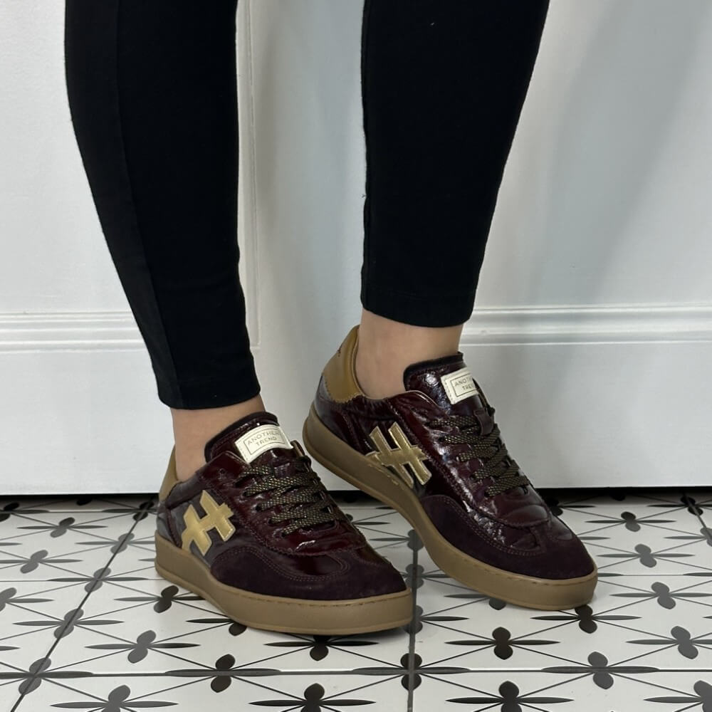 A person wearing Another Trend glossy burgundy leather sneakers with tan suede panels and gold cross-shaped side logos, paired with black leggings, standing on a black-and-white patterned tile floor.