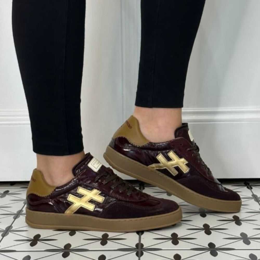 A person wearing Another Trend glossy burgundy leather sneakers with tan suede panels and gold cross-shaped side logos, paired with black leggings, standing on a black-and-white patterned tile floor.