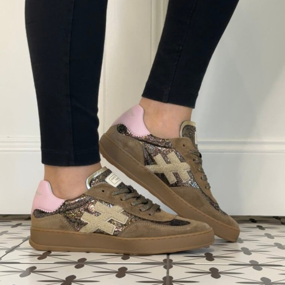 A person wearing black leggings and Another Trend  brown low-top sneakers with pink heel tabs and glittery beige cross logos, standing on a patterned tile floor against a white wall.