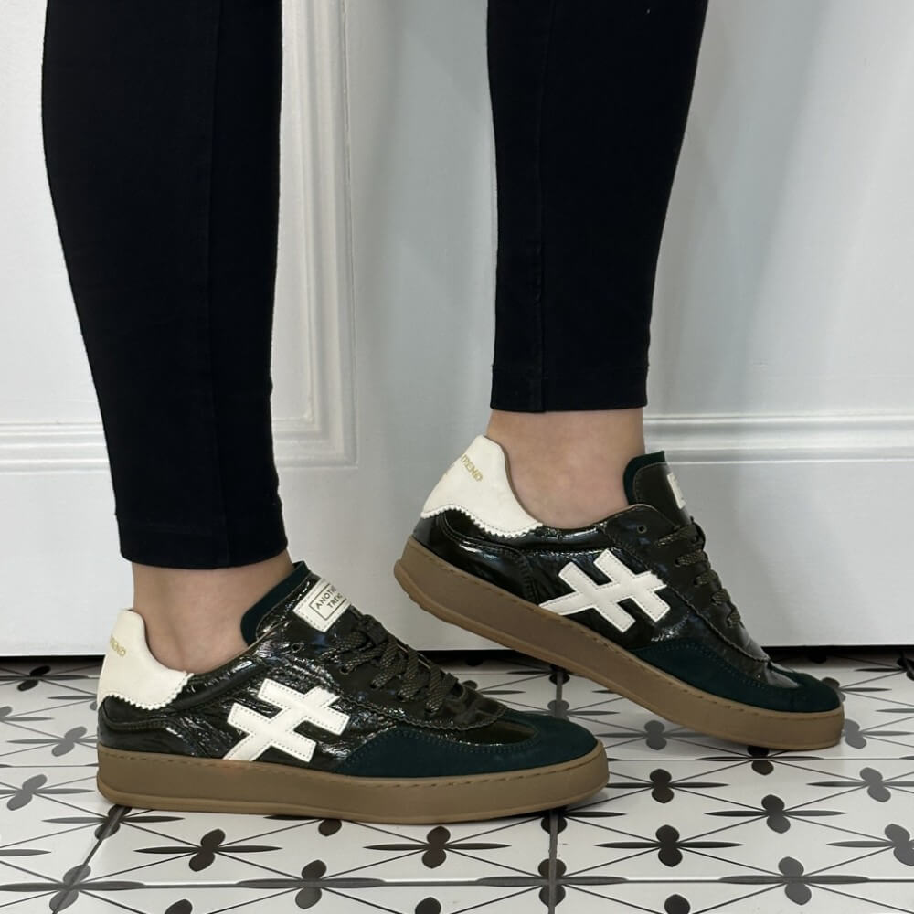 A person wearing black leggings and Another Trend dark green patent leather sneakers with white '#' side stripes, cream heel tabs, and brown gum soles, standing on a black-and-white patterned tile floor against a white panelled wall.