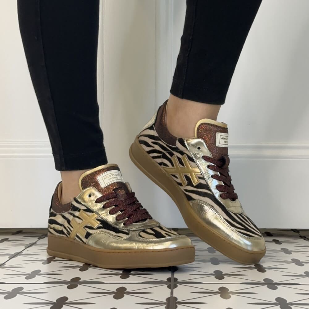 A person wearing black skinny jeans and Another Trend metallic zebra-print sneakers with gold cross logos, standing on a patterned tile floor against a white wall.