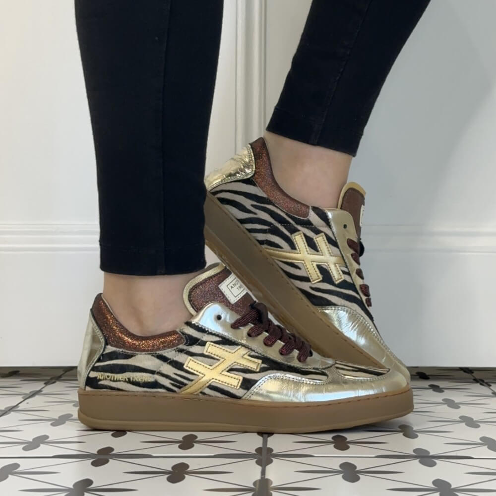 A person wearing black skinny jeans and Another Trend metallic zebra-print sneakers with gold cross logos, standing on a patterned tile floor against a white wall.