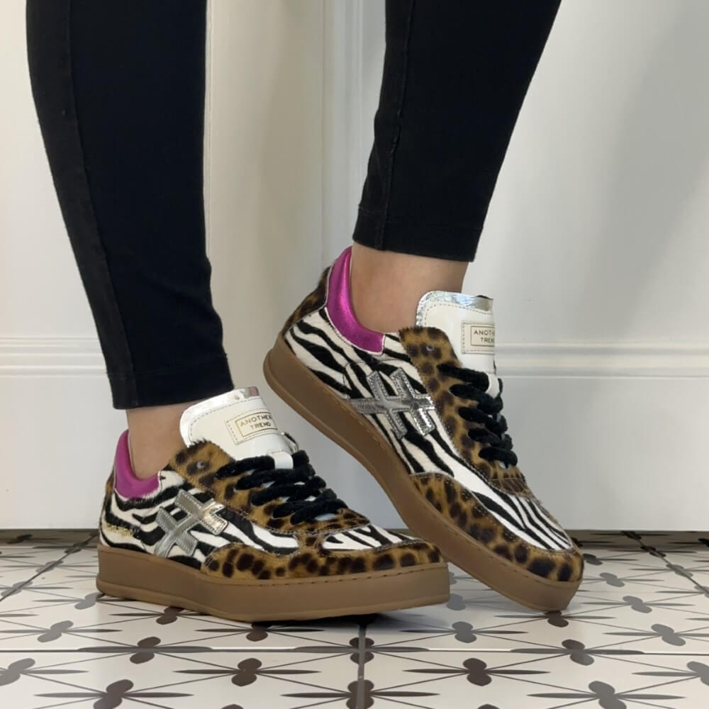 A person wearing black leggings and Another Trend  Leopard and Zebra print low-top sneakers with pink heel tabs and silver cross logos, standing on a patterned tile floor against a white wall.