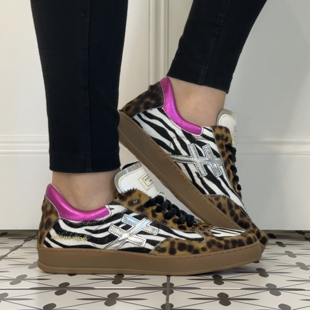 A person wearing black leggings and Another Trend  Leopard and Zebra print low-top sneakers with pink heel tabs and silver cross logos, standing on a patterned tile floor against a white wall.