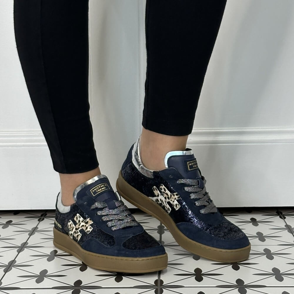 A person wearing Another Trend navy blue glitter sneakers with leopard print overlays and silver accents, paired with black leggings, standing on a black-and-white patterned tile floor.