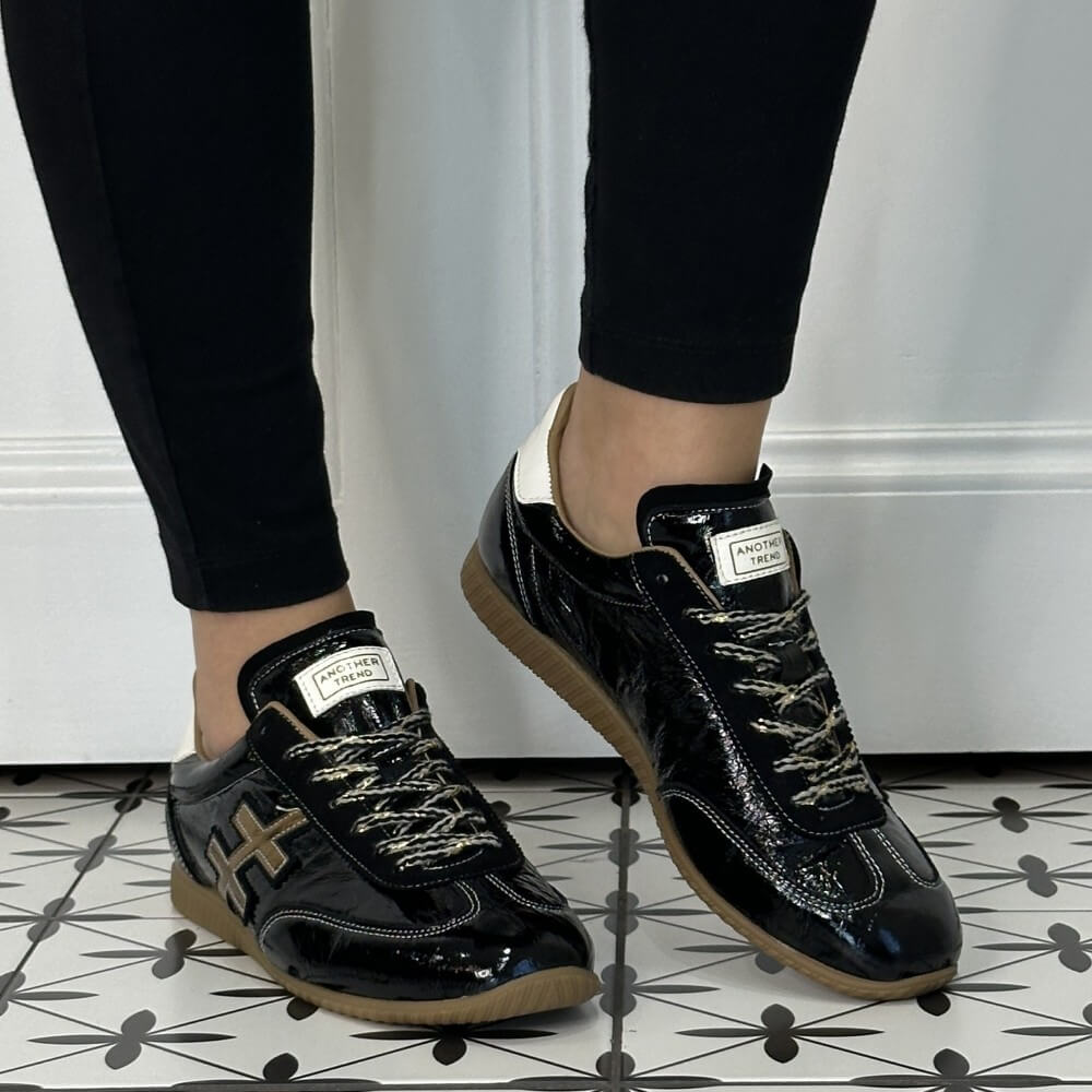 Another Trend Low profile Black patent leather trainers with gold and white accents worn with black jeans on a tiled floor 