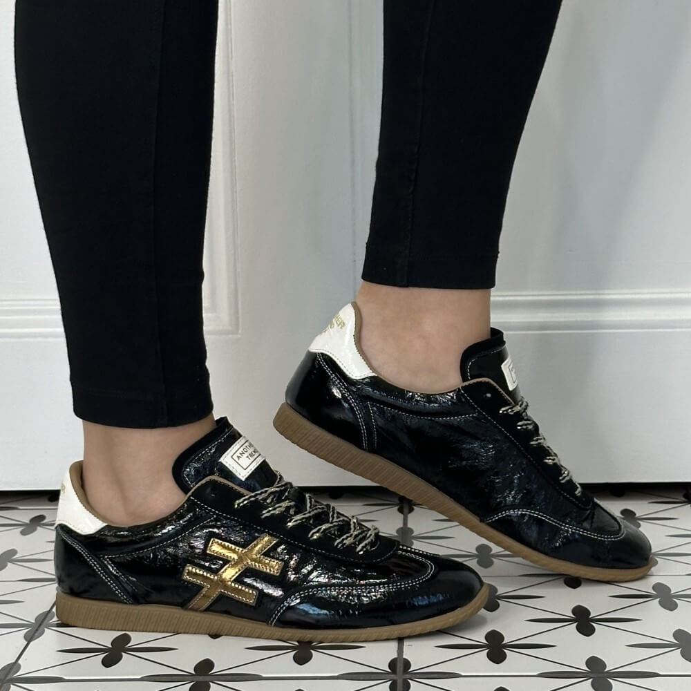 Another Trend Low profile Black patent leather trainers with gold and white accents worn with black jeans on a tiled floor 