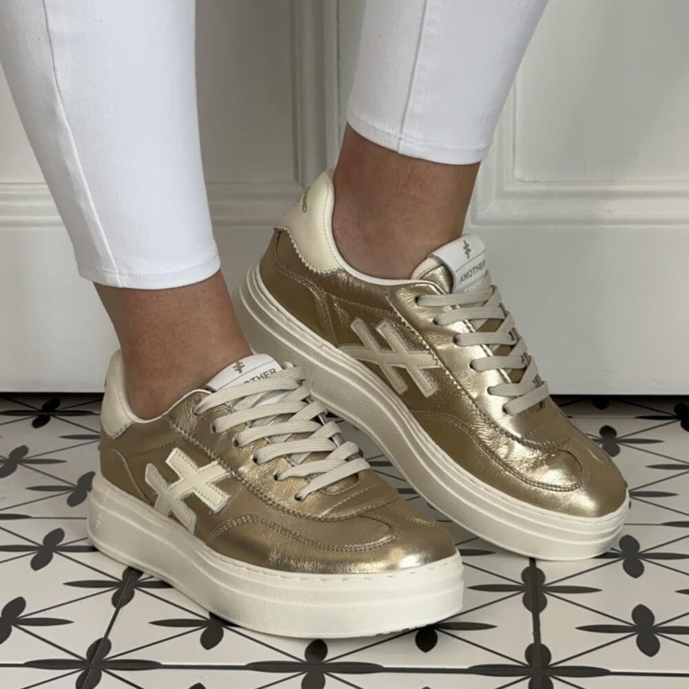 Another Trend Gold sneakers worn with white pants on a patterned floor.