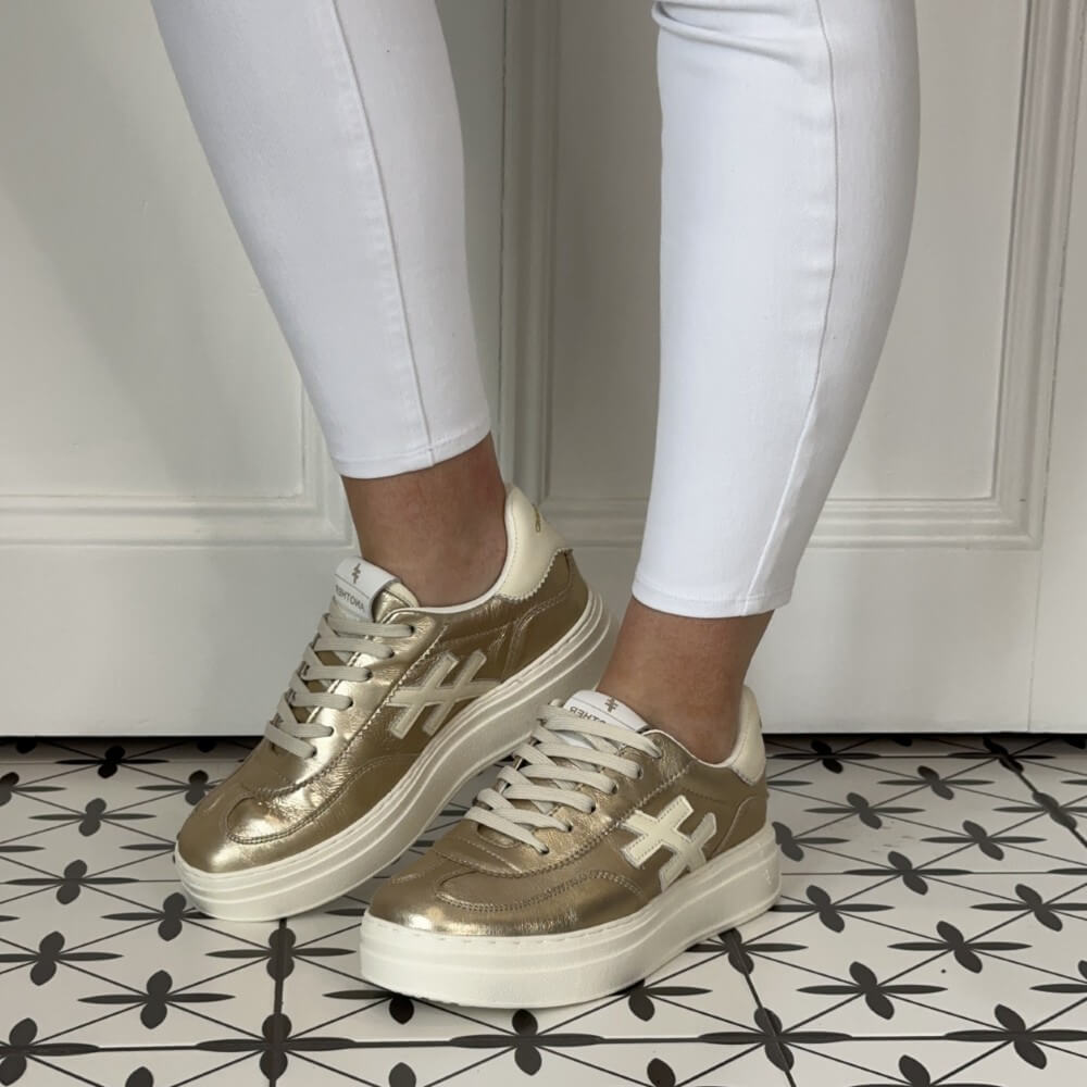 Another Trend Gold sneakers worn with white pants on a patterned floor.